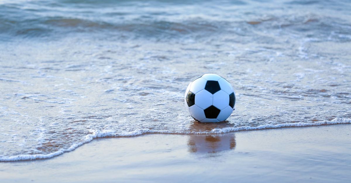 A soccer ball rests on the sandy shore as gentle waves reach it under daylight.